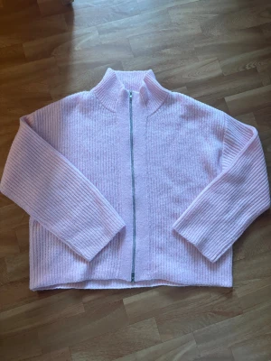 Ljusrosa stickad kofta från 157 - Mysig ljusrosa stickad kofta från lager 157 med rak passform och långärmade ribbade ärmar. Koftan har en hög krage och silverfärgad dragkedja framtill. Perfekt för dig som gillar en chill och oversized look. Materialet är mjukt och ger en skön känsla.