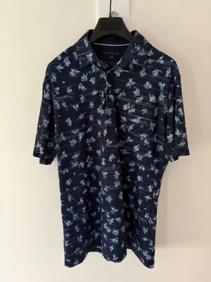 Mörkblå mönstrad golfpiké Galvin Green - Snygg mörkblå golfpiké från Galvin Green med ljusblå blommigt mönster. Pikén har klassisk krage, korta ärmar. Perfekt för dig som gillar en avslappnad men stilren look. Perfekt till golfbanan. Storlek: L passar M. Nypris: 999kr