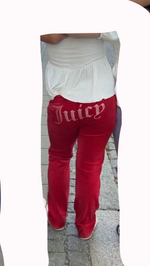 Röda velourbyxor från Juicy Couture - Säljer ett par röda velourbyxor från Juicy Couture med glittrande Juicy-text i strass över bakfickan. Byxorna har en rak passform och normal midja, mjukt material och klassisk Juicy-stil. Perfekta för dig som gillar statement-plagg och comfy vibes. dom saknar snöret annars ingen fel, använda kanske 7 gånger