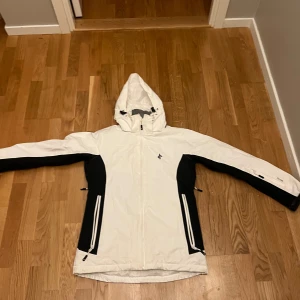 Vit och svart vindjacka Cross Sportswear S - Snygg vit och svart vindjacka från Cross Sportswear i storlek S. Jackan har huva, dragkedja framtill och två stora fickor med dragkedja. Perfekt för kyliga dagar och har sportig look med svarta paneler längs sidor och ärmar.