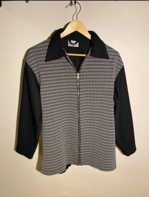 Svart och grå overshirt från Falk collection - Overshirt med svart och grått rutigt mönster framtill, helsvart baksida och ärmar. Jackan har krage, dragkedja och trekvartsärmar. Materialet känns mjukt och lite stretchigt, perfekt för lager-på-lager. Snygg och unik design som sticker ut.