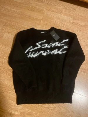 Svart Saint Laurent stickad tröja - Svart stickad tröja från Saint Laurent med vit, handskriven logga över bröstet. Tröjan har rund hals, långa ärmar och ribbade muddar. Materialet är mjukt och fluffigt, perfekt för att hålla värmen. Snygg och lyxig vibe med tydlig designerstil. Använd 1-2 gånger max