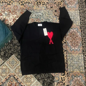 Svart stickad tröja från Ami - Svart stickad tröja från Ami med ett stort rött hjärta och bokstaven A broderat på bröstet. Rund hals och ribbade muddar vid ärmslut och nederkant. Perfekt för dig som gillar stilrena detaljer med en twist.