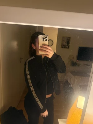 Svart kofta  från Karl Lagerfeld - Svart kofta  från Karl Lagerfeld med vita logoband längs ärmarna. Jackan har dragkedja framtill, ståkrage och raka fickor. Snygg och sportig look som sticker ut med tydlig branding på ärmarna. ÄKTA ALDRIG ANVÄNT  köptes för 2000 kr säljer för 700 kr men pris kan diskuteras 