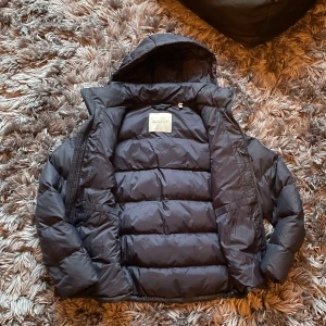 Mörkblå pufferjacka från GANT - Säljer en mörkblå pufferjacka från GANT med huva och dragkedja framtill. Jackan har ett quiltat mönster och är perfekt för kalla dagar. Flera fickor med dragkedja och en stilren look som funkar till de flesta outfits.