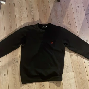 Svart tröja från Polo Ralph Lauren - Svart långärmad tröja från Polo Ralph Lauren med klassisk rund hals och röd broderad logga på bröstet. Tröjan har en clean och stilren look, perfekt för dig som gillar enkel design med en touch av lyx. Materialet känns mjukt och har en lätt stretch.