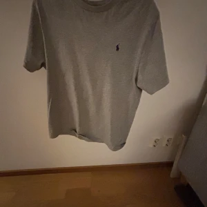 Grå t-shirt från Polo Ralph Lauren - Snygg grå t-shirt från Polo Ralph Lauren med klassisk passform och korta ärmar. Diskret logga broderad på bröstet i mörkblått. Perfekt basic-plagg med mjukt bomullsmaterial och rund hals.