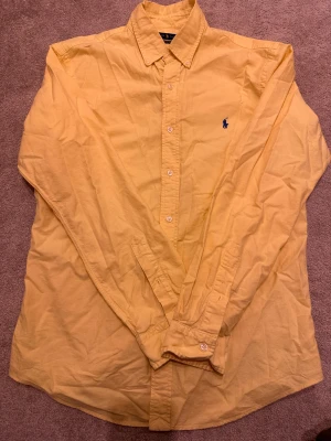 Gul skjorta från Ralph Lauren - Klassisk gul skjorta från Ralph Lauren i slim fit-modell. Skjortan har långa ärmar, knappar framtill och en broderad blå logga på bröstet. Tillverkad i bomull och har en stilren, tidlös look med button-down krage.