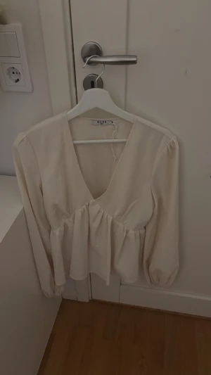 Beige v-ringad blus från NA-KD - Säljer en beige blus från NA-KD med v-ringning och volangdetalj under bysten. Blusen har långa ärmar med lätt puff och är tillverkad i ett mjukt, vävt material. Perfekt för dig som gillar en feminin och chill stil.