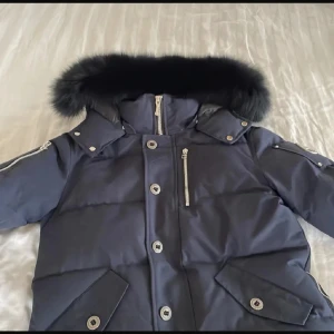 Mörkblå dunjacka Moose Knuckles - Snygg mörkblå dunjacka från Moose Knuckles med päls på huvan, silverfärgade detaljer och flera fickor med dragkedja och knappar. Jackan har lång ärm och quiltad design, perfekt för kalla dagar. Märkets ikoniska logga på ärmen. Pris går att diskutera vid snabb affär 