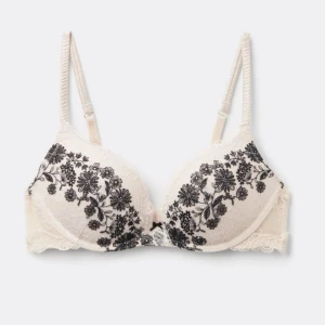 Beige intimissimi bh med svart blommig brodyr - Supersnygg bh i beige spets med svarta blommiga broderier över kuporna. Justerbara axelband och hakknäppning bak. Kuporna är lätt vadderade och har en liten rosett framtill. Perfekt för dig som gillar romantiska detaljer och stilren design.