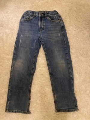 Blå jeans från Lindex, storlek 140 - Snygga blå jeans från Lindex i rak modell med slitna detaljer och fransade benslut. Klassisk femficksdesign och resår i midjan för extra komfort. Perfekta för dig som gillar en avslappnad och cool stil.