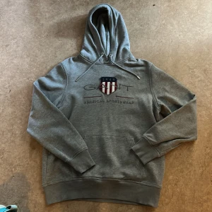 Grå hoodie från GANT med brodyr - Säljer en grå hoodie från GANT i storlek M. Tröjan har broderad logga och sköld på bröstet, samt texten 'American Sportswear'. Klassisk känguruficka framtill och huva. Perfekt för chill dagar eller när du vill ha en clean streetstyle. Har andvänt typ 10 gånger 