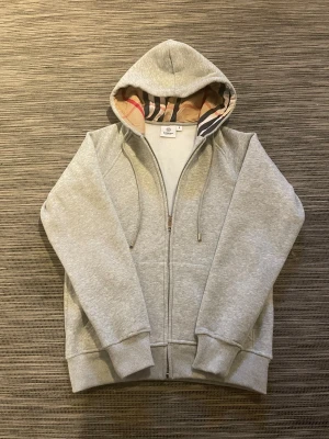 Grå hoodie från Burberry med mönstrad huva - Säljer en snygg Burberry hoodie! Hoodien är i mycket bra skick och har inga defekter. Mjukt material inuti tröjan vilket gör den skön och varm. Om ni har några frågor så är de bara att höra av er!