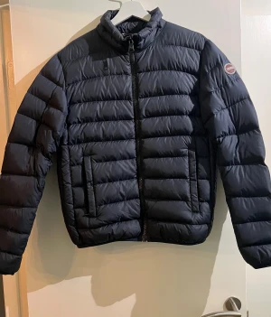 Svart pufferjacka från Colmar - Snygg marinblå pufferjacka från Colmar med hög krage och raka, horisontella sömmar. Jackan har dragkedja framtill, två fickor och diskret logga på ärmen. Perfekt för kyliga dagar och ger en clean, modern look.