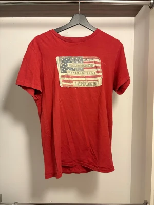 Röd t-shirt från Ralph Lauren - Snygg röd t-shirt från Ralph Lauren med coolt amerikanskt flaggtryck och text på bröstet. Klassisk rund hals och korta ärmar. Perfekt för dig som gillar streetstyle och vill sticka ut med en ikonisk look.