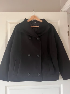 Svart skepparkavaj med bred krage - Aldrig använd.Säljer en svart skepparkavaj med bred krage och dubbelknäppning. Jackan har en loose passform, långa ärmar och två sidofickor. Materialet är ullblandning som ger en mjuk och varm känsla. Perfekt för dig som gillar stilrena och klassiska vibes.