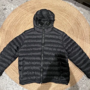 Marinblå pufferjacka från C.P. Company - Säljer en svart pufferjacka från C.P. Company med ikoniska goggles i huvan. Jackan har Pro-Tek superflex tekniskt ytterlager, dragkedja framtill och två sidofickor med dragkedja. Perfekt för dig som vill ha en unik och funktionell jacka med streetkänsla. Jackan är perfekt nu till vintern för den som både vill hålla sig varm och gå runt med stil. 