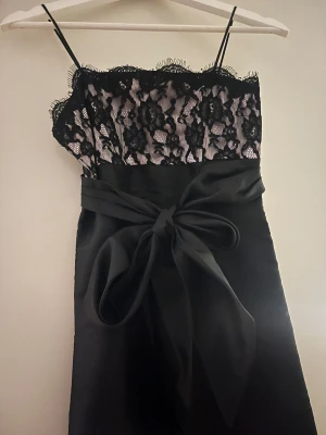 Svart spetsklänning med rosett framtill - Supersnygg svart klänning med smala axelband, spets med blommönster och stor rosett framtill, med lapp kvar. Det står Stolek S/M men jag skulle säga att den är i Stolek Xs möjligen en liten S.