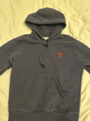 Mörkblå hoodie från Ami Paris - Säljer en mörkblå hoodie från Ami Paris med klassisk känguruficka och snörning i huvan. På bröstet finns Ami:s ikoniska röda hjärta med A broderat. Tröjan är i mjuk bomull och har en relaxed passform, perfekt för chill dagar. Är lite små smutsig på bild men du får den ny tvättad hem