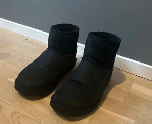 Svarta UGGS herr - Klassiska svarta UGG boots i mjuk mocka med rund tå och platt sula. Insidan är fodrad med mysig päls för extra värme. Perfekta för kalla dagar och har en enkel, stilren look med UGG-logga bak på hälen.
