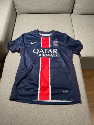PSG matchtröja Nike Champions of Europe - Paris Saint-Germain fotbollströja i mörkblått med röd och vit rand på framsidan, PSG-emblem och guldstjärna på bröstet. Tryck med Qatar Airways och Champions of Europe 25 på ryggen. Tillverkad i lätt och ventilerande polyester.