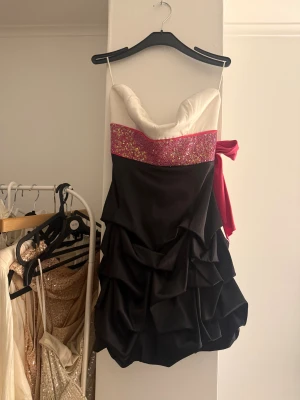 Glittrig kort klänning med volanger - Superfin kort klänning med vit överdel, glittrigt rosa parti med paljetter och stor rosa rosett i ryggen. Kjolen är svart med flera lager volanger som ger en cool och festlig look. 