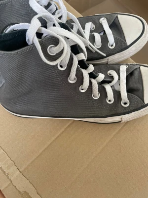 Grå Converse Chuck Taylor All Star - Klassiska höga Converse Chuck Taylor All Star i grå canvas med vita snören och gummisula. Skorna har rund tå, vita detaljer och den ikoniska loggan på sidan. Perfekta för en avslappnad och cool stil.