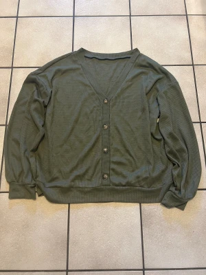 Olivgrön ribbad tröja  med dekorativa knappar - Olivgrön tröja med ribbad struktur och v-ringad hals. Tröjan har fem knappar framtill och långa ärmar med muddar. Materialet känns mjukt och lätt, perfekt för lager-på-lager. Enkel och stilren design som funkar till många olika looks.