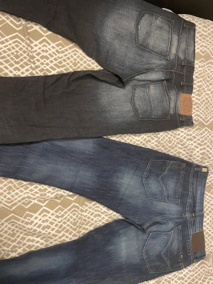 Replay Anbass och Grover jeans blå - Två par snygga Replay jeans, ena modellen Anbass och den andra Grover. Båda är blå med slitningar och har klassisk femficksdesign. Jeansen har raka ben och normal passform, tillverkade i slitstarkt denim med Replay-logga på bakfickan.