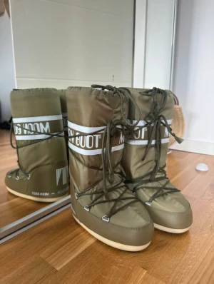 Olivgröna Moon Boots med snörning - Säljer ett par olivgröna Moon Boots med vit logga och snörning framtill. Skorna har en hög och vadderad design med platt sula och rund tå. Perfekta för kalla dagar och vinterns alla äventyr. Utsidan är i syntetmaterial och insidan är mjukt fodrad. Använd bara 1 gång.