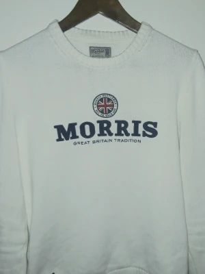 Vit stickad tröja från Morris - Snygg vit stickad tröja från Morris med broderad logga och brittisk flagga på bröstet. Rund hals och ribbade muddar ger en klassisk look. Perfekt för dig som gillar stilrena och tidlösa plagg med en touch av brittisk tradition.