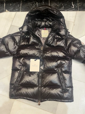 Moncler Maya  - Säljer en svart pufferjacka från Moncler med glansig finish och huva. Jackan har två snedställda fickor med dragkedja, Moncler-logga på ärmen och quiltad design. Perfekt för dig som vill ha en stilren och varm jacka med street vibe.