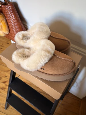 UGG Disquette platform slippers - Supermysiga slip-in skor från UGG i beige mocka med fluffig päls runt öppningen. De har en rund tå och platt sula, perfekt för chill dagar hemma. Snyggt UGG-logo på sidan och extra mjuk insida för maxad komfort.