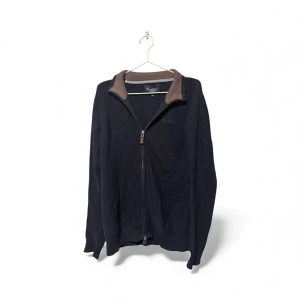 Svart kofta med dragkedja från Bondelid - Svart ribbad kofta från Bondelid i storlek M. Koftan har en brun krage med kontrastdetalj och hel dragkedja framtill. Perfekt för dig som gillar klassisk stil med en twist. Materialet är stickat och känns mjukt mot huden.