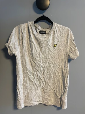Grå t-shirt från Lyle & Scott - Snygg grå t-shirt från Lyle & Scott med klassisk rund hals och korta ärmar. På bröstet sitter den ikoniska gula fågel-loggan som ger en stilren touch. Tillverkad i mjuk bomull för skön komfort hela dagen.
