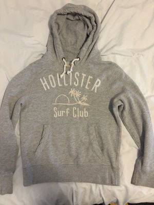 Vintage grå Hollister Surf Club Hoodie - Grå hoodie från Hollister med broderad text 'Surf Club' och palmmotiv på bröstet. Tröjan har huva med snörning, känguruficka och ribbade muddar vid ärmslut och nederkant. Perfekt för chill dagar och sköna häng. Liten defekt vid ärmen syns på bild 3 