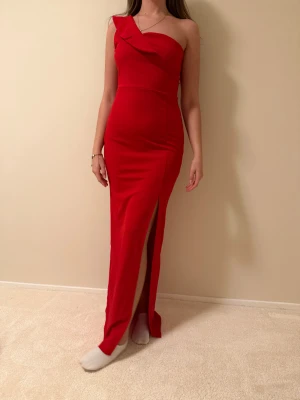 Röd långklänning med slits och oneshoulder - Säljer en snygg röd långklänning med en axel och cool draperad detalj upptill. Klänningen har en figurnära passform och en hög slits framtill som ger en extra touch. Helt ny prislappen är på.