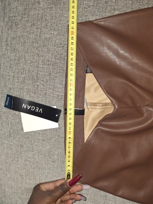 Bruna veganska skinnbyxor hm - Snygga bruna veganska skinnbyxor från hm med slits vid benslut. Byxorna har en rak passform och hög midja, vilket ger en trendig siluett. Materialet är mjukt syntetiskt skinn med beige insida. Perfekt för dig som vill ha en cool och modern look.