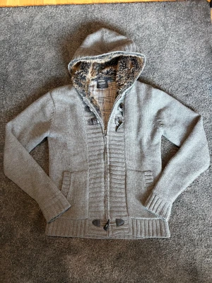 Grå stickad cardigan jacka Pull&Bear - Grå stickad cardigan jacka från Pull&Bear med mysig fuskpäls i luvan och rutigt innerfoder. Jackan har dragkedja, snygga knappar i trä-look och två fickor framtill. Perfekt för kalla dagar och har en chill, urban vibe.