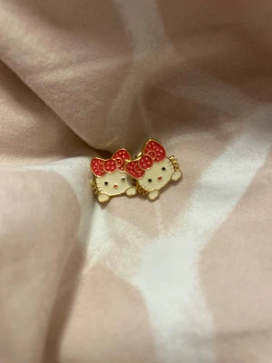 Hello Kitty örhängen med röd rosett - Supersöta Hello Kitty örhängen i form av kattens ansikte med röd prickig rosett. Örhängena är i guldtonad metall med vita och röda detaljer, perfekta för dig som älskar kawaii och vill sticka ut lite extra.