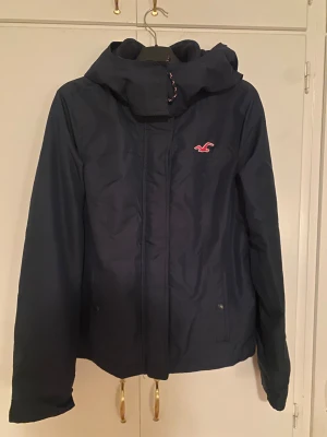 Mörkblå vinterjacka från Hollister M - Säljer en mörkblå vinterjacka från Hollister med huva, dragkedja och knappar. Jackan har mjukt rosa innerfoder och Hollister-logga på bröstet. Praktiska fickor framtill. 