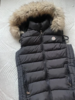 Svart dunväst från Moncler med pälskant - Svart dunväst från Moncler med fluffig pälskantad huva och quiltad design. Västen har dragkedja framtill, två fickor med dragkedja och Moncler-logga på bröstet. Perfekt för kalla dagar när du vill hålla stilen.