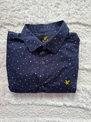 Mörkblå prickig skjorta Lyle & Scott - Mörkblå skjorta från Lyle & Scott med vita prickar och klassisk krage. Skjortan har långa ärmar, knäppning framtill och den ikoniska gula örnloggan broderad på bröstet. Tillverkad i mjuk bomull för en skön känsla.