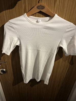 Vit ribbad t-shirt från C.W. - Fräsch vit t-shirt från C.W. i storlek XS. Modellen är ribbad med rund hals och korta ärmar, vilket ger en clean och stilren vibe. Perfekt att matcha med jeans eller shorts för en enkel look. Materialet känns mjukt och skönt mot huden.