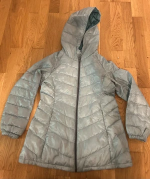 Ljusblå dunjacka med huva från Uniqlo - Säljer en tunnare ljusblå dunjacka från Uniqlo med huva och hel dragkedja framtill. Jackan är quiltad och har elastiska muddar vid ärmsluten. Perfekt för kyliga dagar och enkel att matcha med olika outfits.