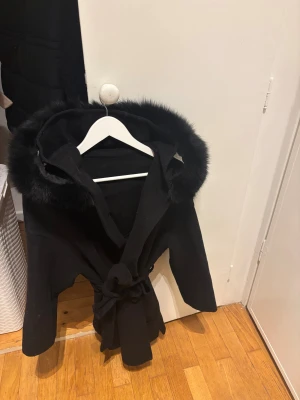 Svart kappa med päls och huva - Stilren svart kappa med stor huva kantad av mjuk fuskpäls. Knyts i midjan med bälte för en snygg siluett. Perfekt för kalla dagar när du vill känna dig både varm och trendig.