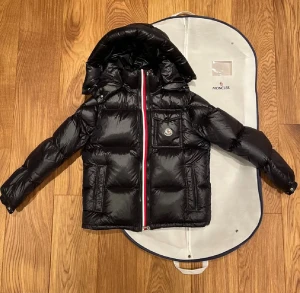 Svart Moncler pufferjacka med huva - KVITTO FINNS 🧾Snygg svart pufferjacka från Moncler med glansig finish, huva och klassisk logga på bröstet. Jackan har en dragkedja med röd, vit och blå rand längs öppningen samt praktiska fickor. Perfekt för kalla dagar när du vill hålla stilen. 