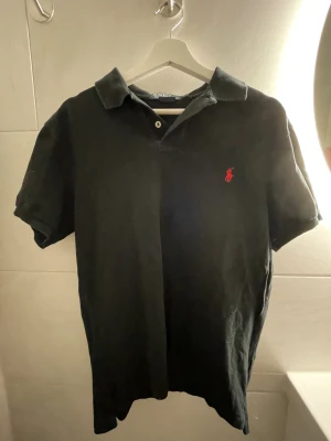 Svart pikétröja från Polo Ralph Lauren - Klassisk svart pikétröja från Polo Ralph Lauren med röd broderad logga på bröstet. Tröjan har korta ärmar, krage och knappar vid halsen. Tillverkad i mjuk bomull som känns skön mot huden. Slim fit 