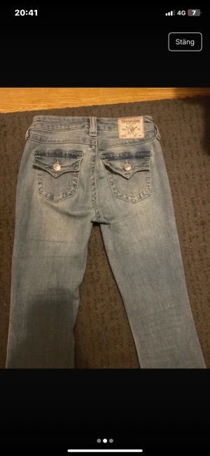 True Religion ljusblå jeans med fickdetaljer - Säljer ett par ljusblå jeans från True Religion med fickor bak, silverfärgade knappar. Modellen har klassisk femficksdesign och ett märke på linningen.